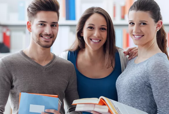 Zwei Studentinnen und ein Student schauen lachend in die Kamera und halten ihre Bücher in der Hand