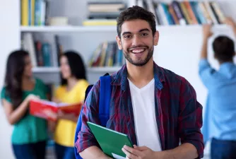 Im Vordergrund steht ein Student mit Buch und lacht in die Kamera. Im Hintergrund sind weitere Studenten am lernen