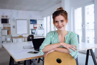 Eine Frau sitzt im Büro und lacht in die Kamera. Im Hintergrund liegt ein Laptop und ein Handy auf dem Tisch