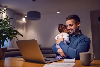 Ein Mann sitzt mit Baby im Arm vor einem Laptop