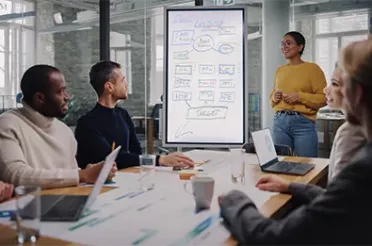 Mehrere Personen sitzen im Meetingraum mit Laptops. Im Hintergrund ist ein Whiteboard