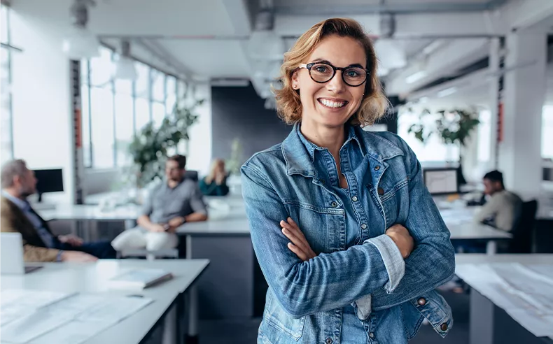 Eine Führungskraft steht im Büro und lächelt mit verschränkten Armen in die Kamera