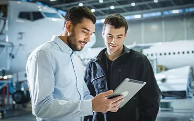 Zwei Männer stehen in einer Halle mit Flugzeugen und schauen auf ein Tablet