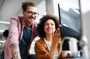 Ein Mann und eine Frau sitzen im Büro lachend vor einem Bildschirm. Die Frau zeigt auf den Bildschirm