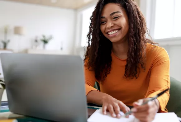 Eine Frau sitzt am Schreibtisch und schaut lachend in ihren Laptop